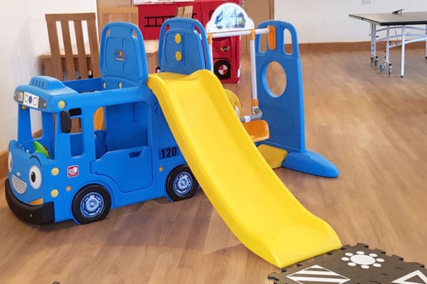 blue children's play bus with yellow slide on a wooden floor with red fireman's truck soft wendy house in background