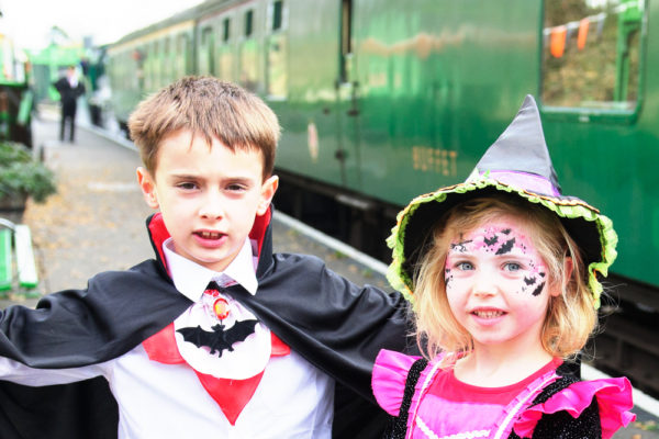 A young boy dressed as Dracula, and a young girl dress in a pink and black witches costume standing on the platform with a green train behind them at The Watercress Line