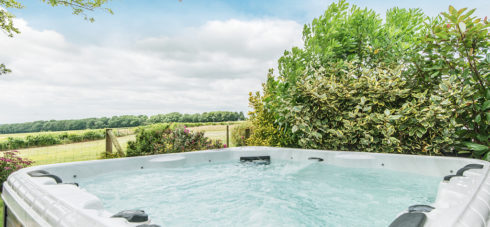 Bubbling hot tub with water jets with tall hedge next to it and views across green fields on a summer day with blue sky